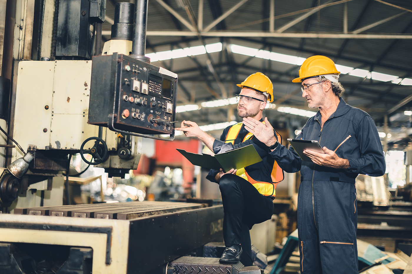Two factory workers wearing yellow hard hats and safety glasses operating and inspecting machinery in a manufacturing facility, one holding a clipboard and the other pointing at machine controls.