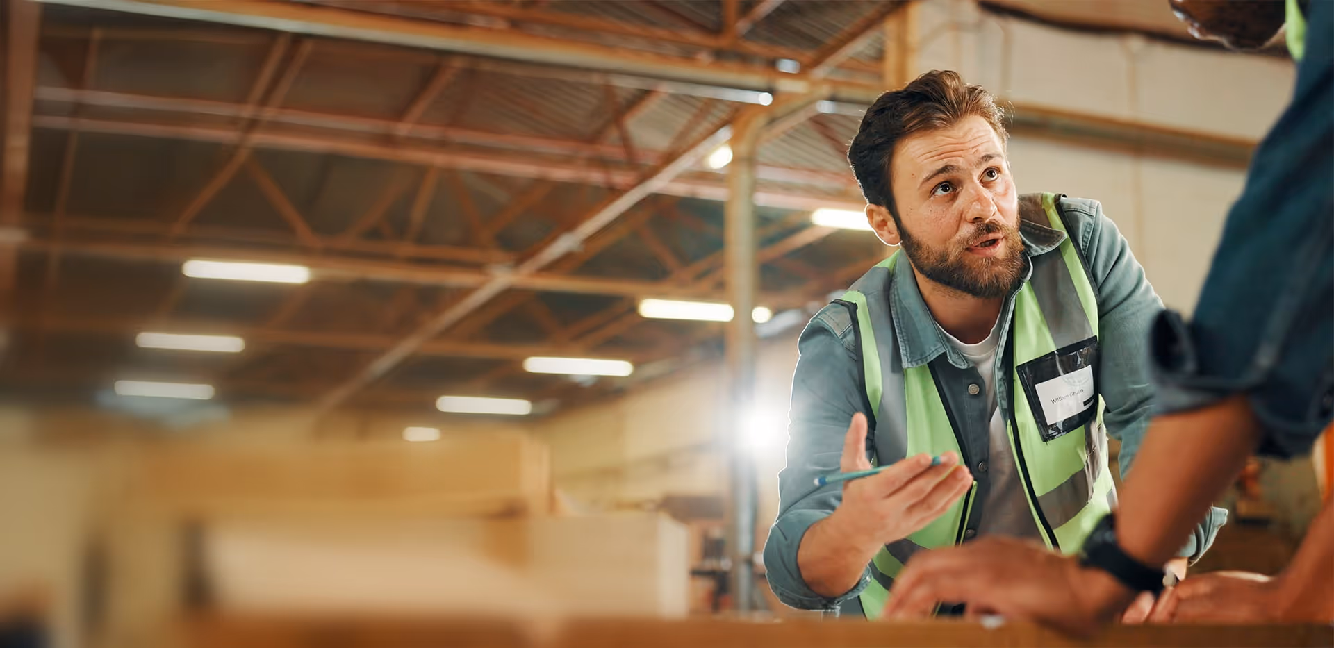 Man wearing a high-visibility vest engaging in a discussion with a colleague inside a warehouse.