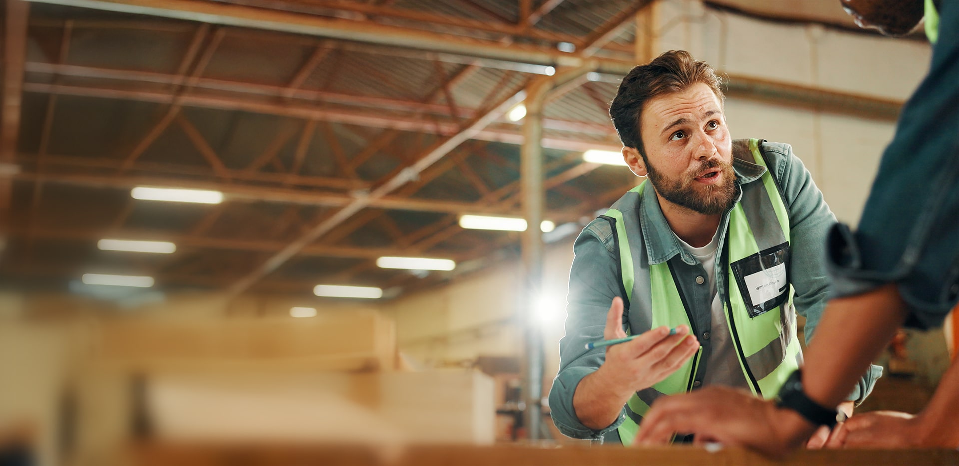 Man wearing a high-visibility vest engaging in a discussion with a colleague inside a warehouse.