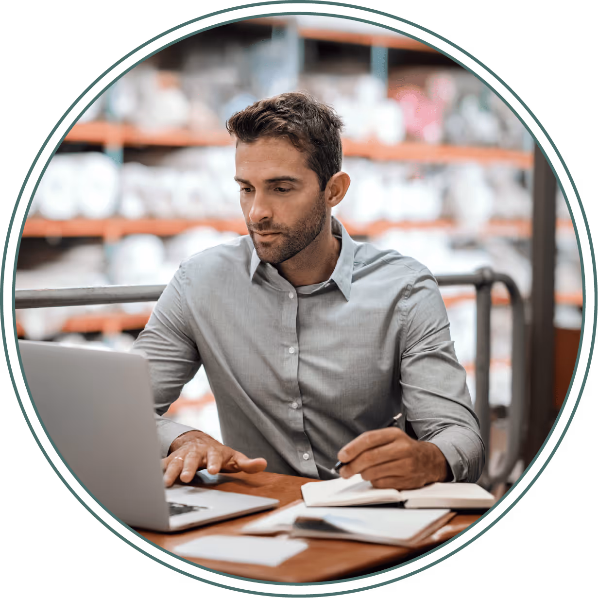 Man in a light grey shirt working on a laptop and writing in a notebook at a wooden table in an industrial setting.
