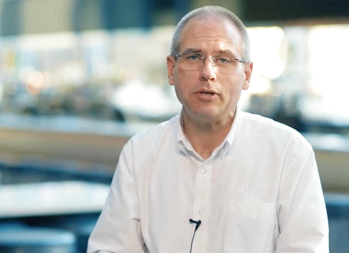 Middle-aged man wearing glasses and a white shirt with AME logo speaking in a blurred indoor setting.