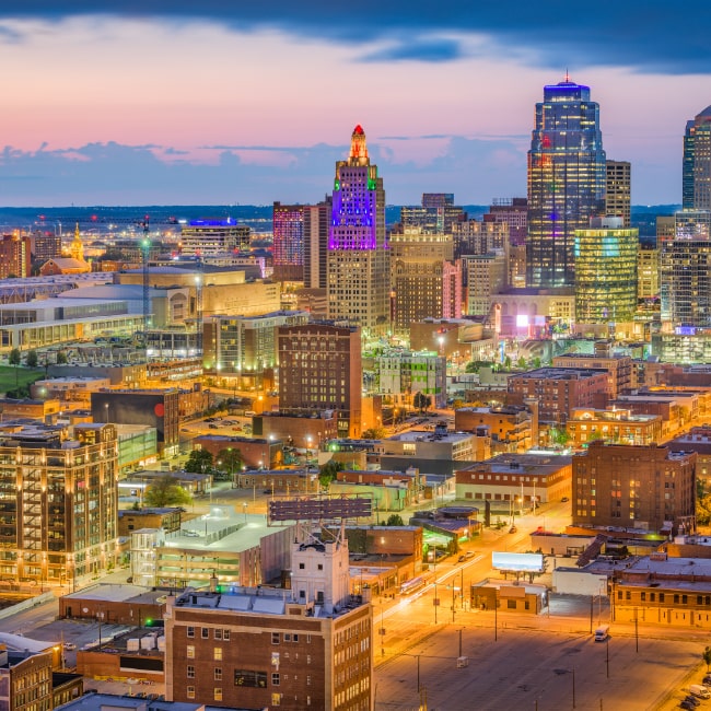 City skyline at dusk with illuminated buildings and streets under a partly cloudy sky.