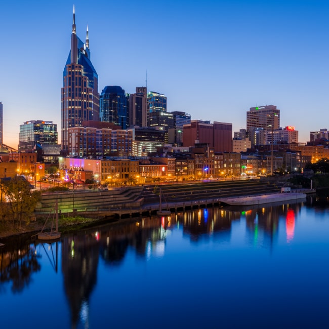Skyline of Nashville at twilight with illuminated buildings reflecting on the calm river.