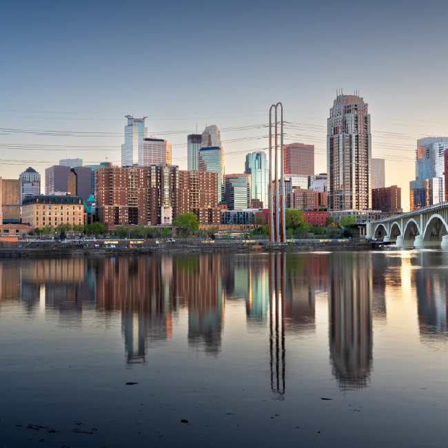 City skyline at sunset reflecting on calm river water with a bridge on the right side.