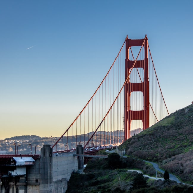Golden Gate Bridge with red towers and suspension cables against a clear blue sky, with green hills in the foreground.