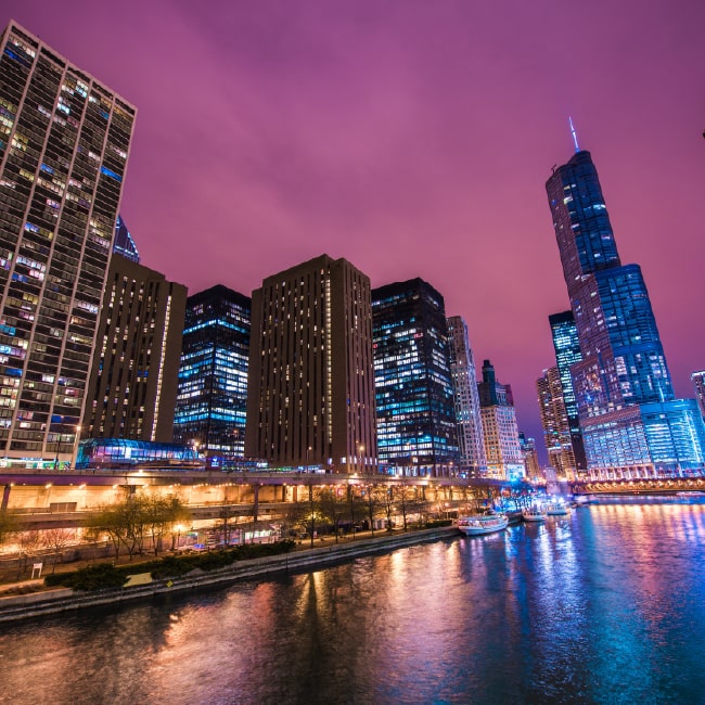 Night cityscape of illuminated skyscrapers along a river with purple sky reflecting on the water.