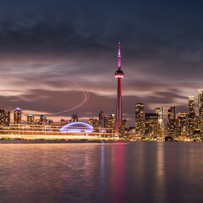 Toronto skyline at dusk with CN Tower lit in red and city lights reflecting on the water.