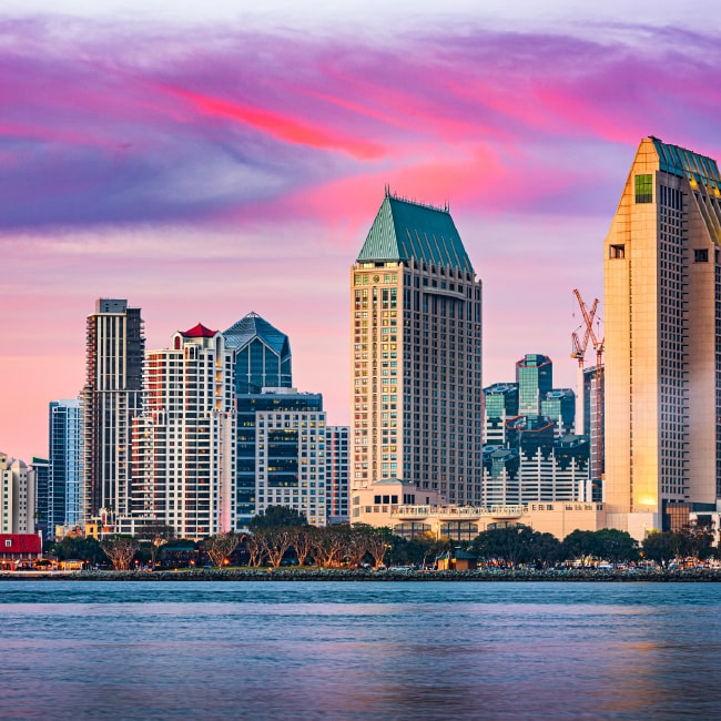 San Diego skyline at sunset with colorful pink and purple clouds over modern high-rise buildings by the waterfront.