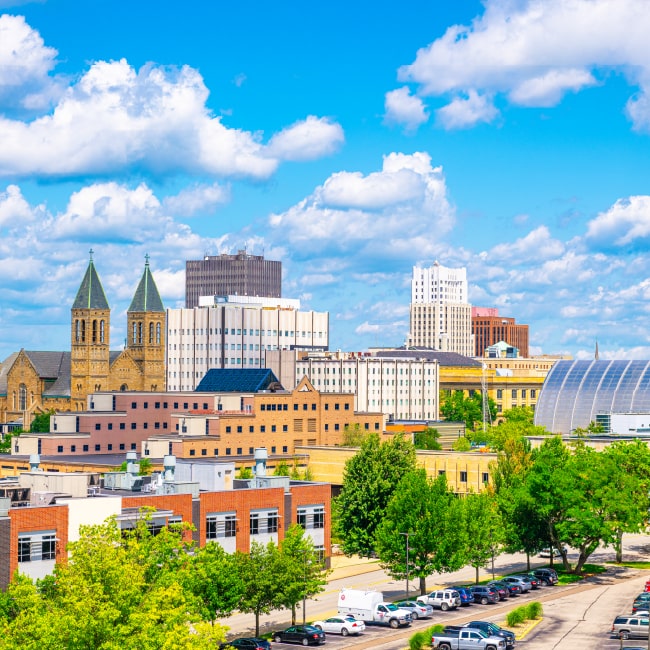 Cityscape with a mix of historic and modern buildings under a bright blue sky with scattered clouds, surrounded by green trees and parked cars.