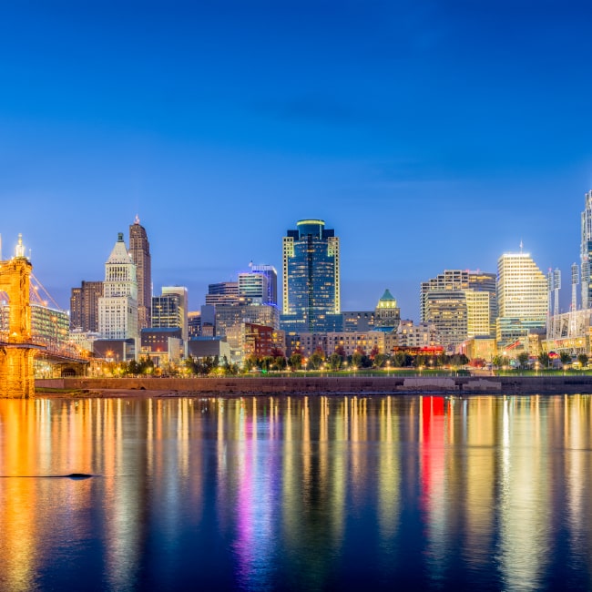 City skyline at dusk with illuminated buildings and bridge reflecting on calm river water.