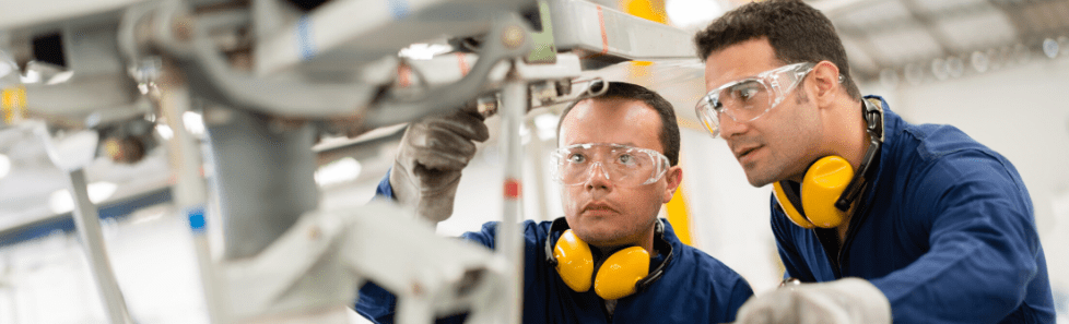 Two male workers wearing safety goggles, gloves, and yellow ear protection inspecting machinery in an industrial setting.