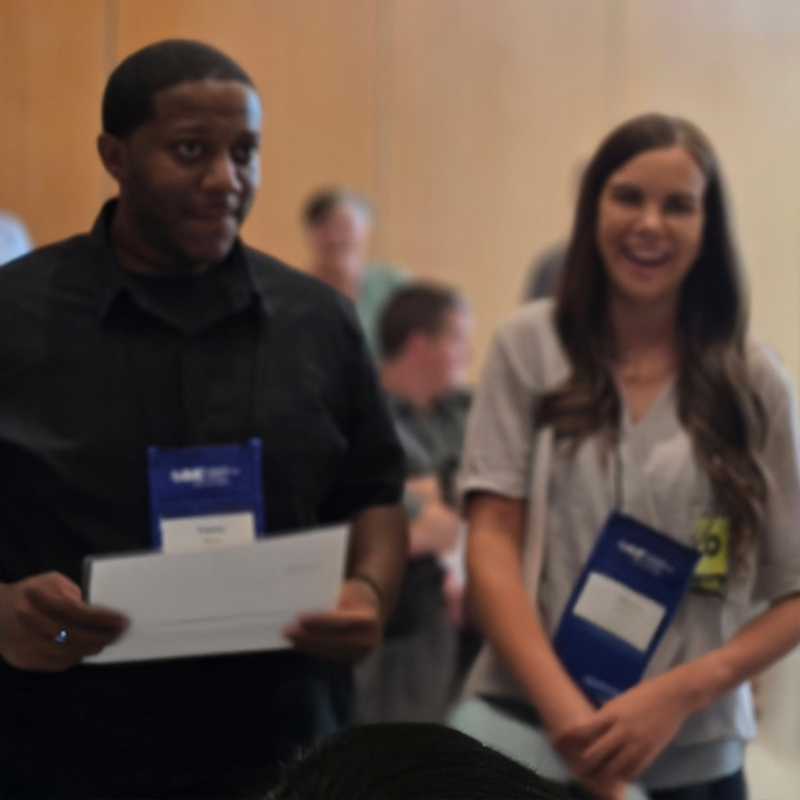 Two people standing and smiling at an indoor event, wearing blue conference badges.