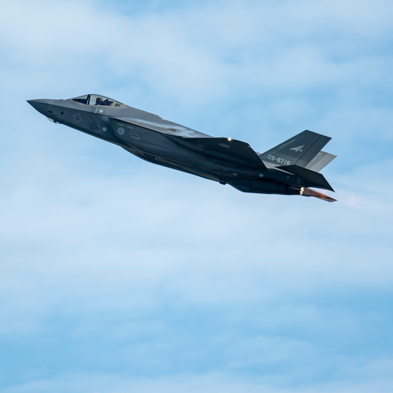 Gray stealth fighter jet climbing against a blue sky with afterburners engaged.