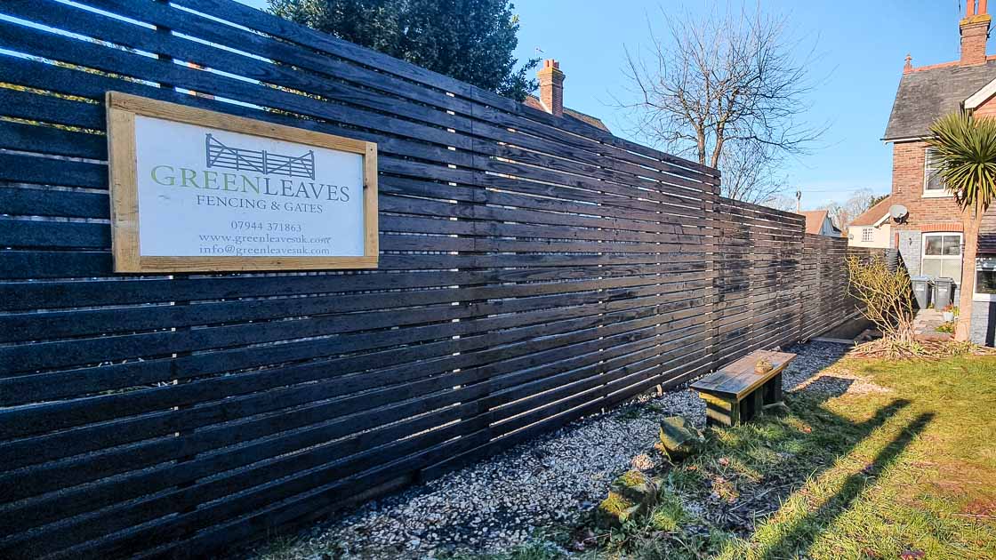 Modern horizontal slatted fence painted black, installed along a sunny garden boundary in East Grinstead.