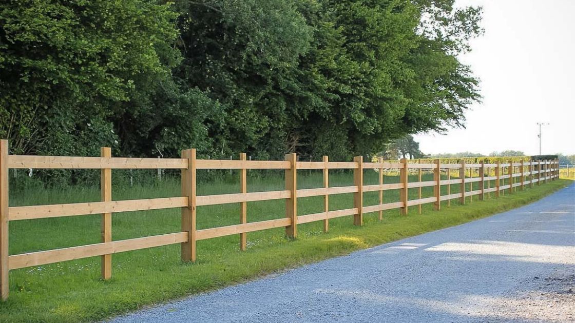 Timber post and rail fencing running beside a country road and open field under a line of trees.
