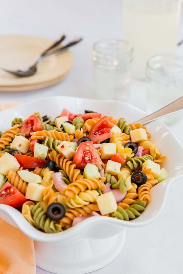 White bowl filled with tri-color rotini pasta salad with diced tomatoes, black olives, cucumber, and cheese cubes.