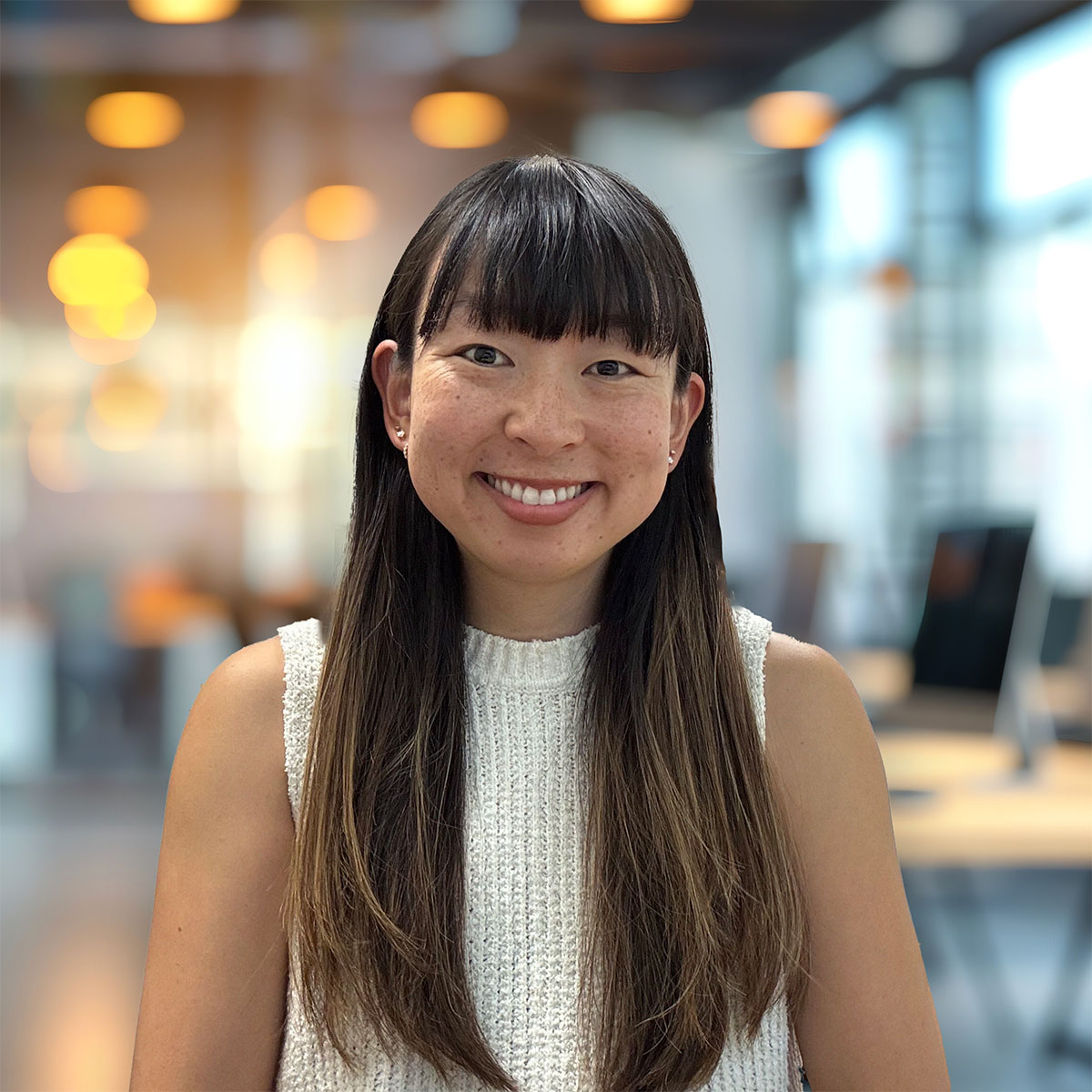 Head-and-shoulders portrait of a smiling woman with long dark hair and bangs, wearing a white knit sleeveless top in a modern office with warm blurred lights.