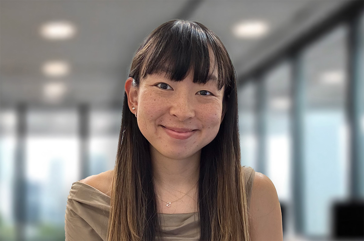 Head-and-shoulders portrait of a smiling woman with long straight hair and bangs, wearing a draped taupe top and small necklace in a bright, modern office with blurred windows.