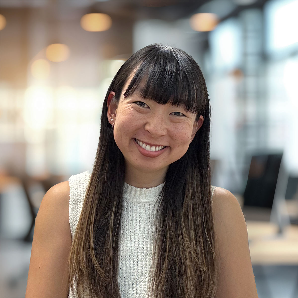 Head-and-shoulders portrait of a smiling woman with long dark hair and bangs, wearing a white knit sleeveless top, in a modern office with warm blurred lights.