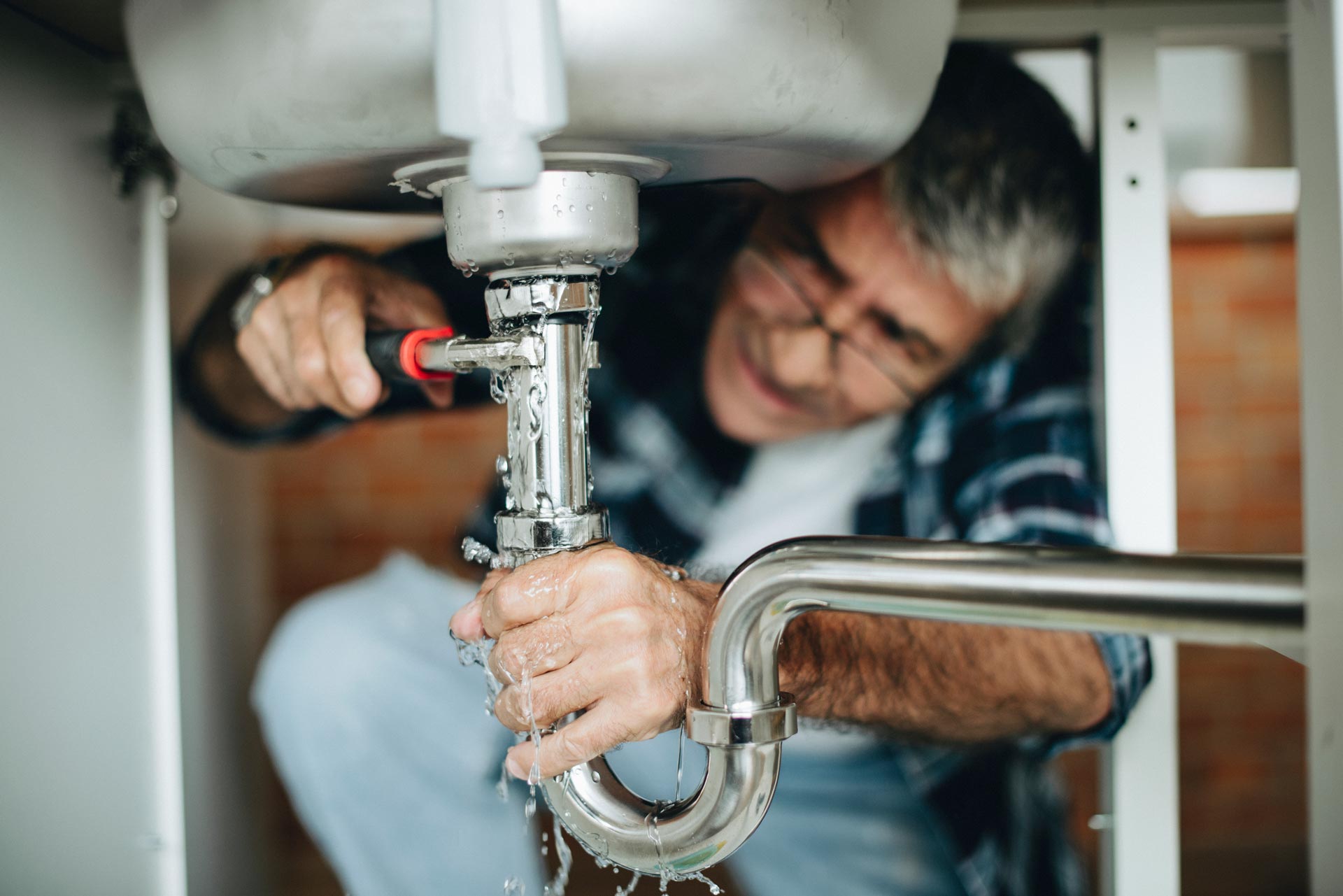 A man using a wrench to fix a leaking kitchen sink pipe under the sink.