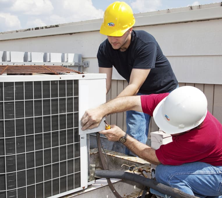 Two technicians wearing hard hats installing or repairing an outdoor air conditioning unit on a rooftop.