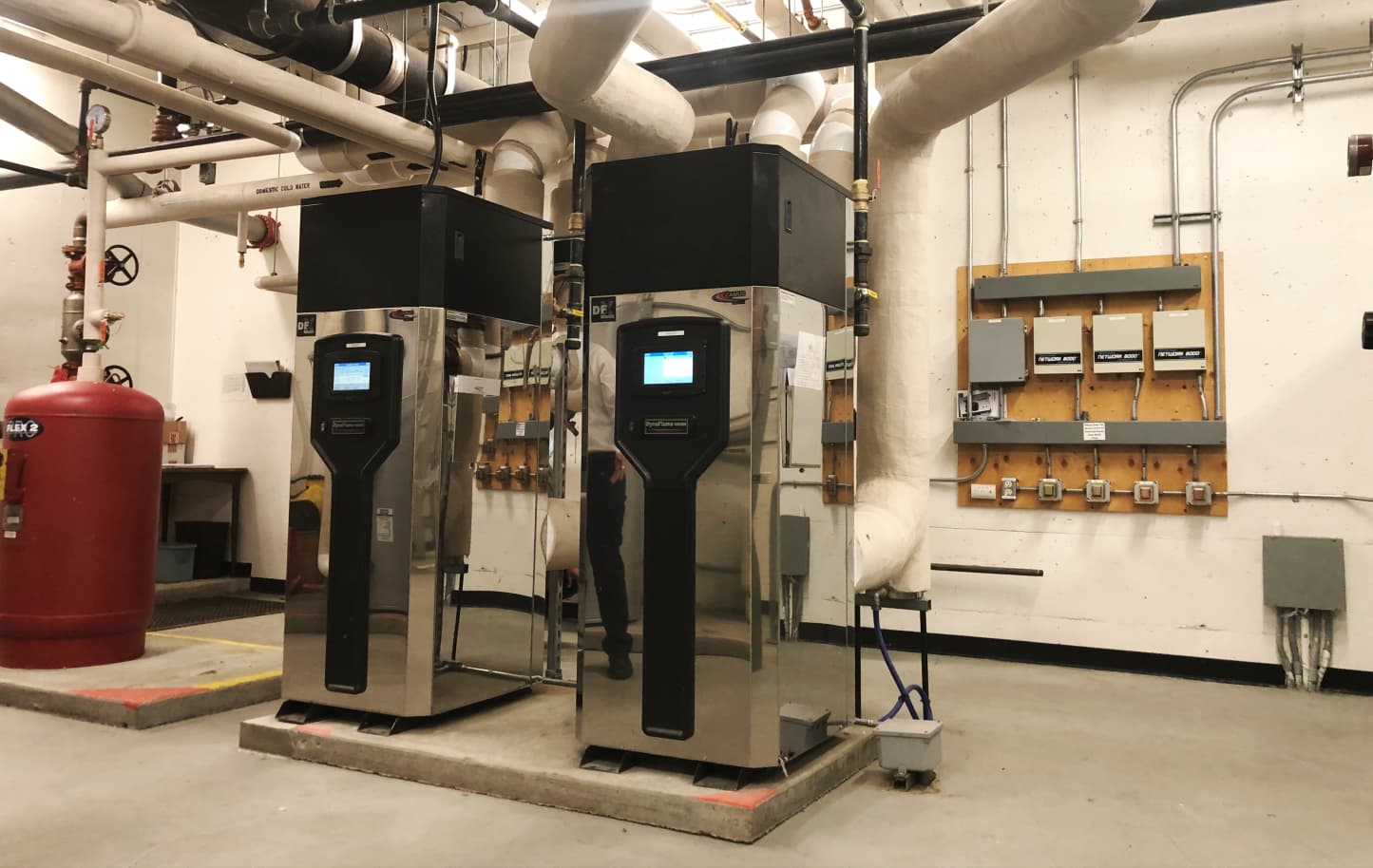 Two large industrial HVAC units with digital control panels installed in a mechanical room with white insulated pipes and a red pressure tank.