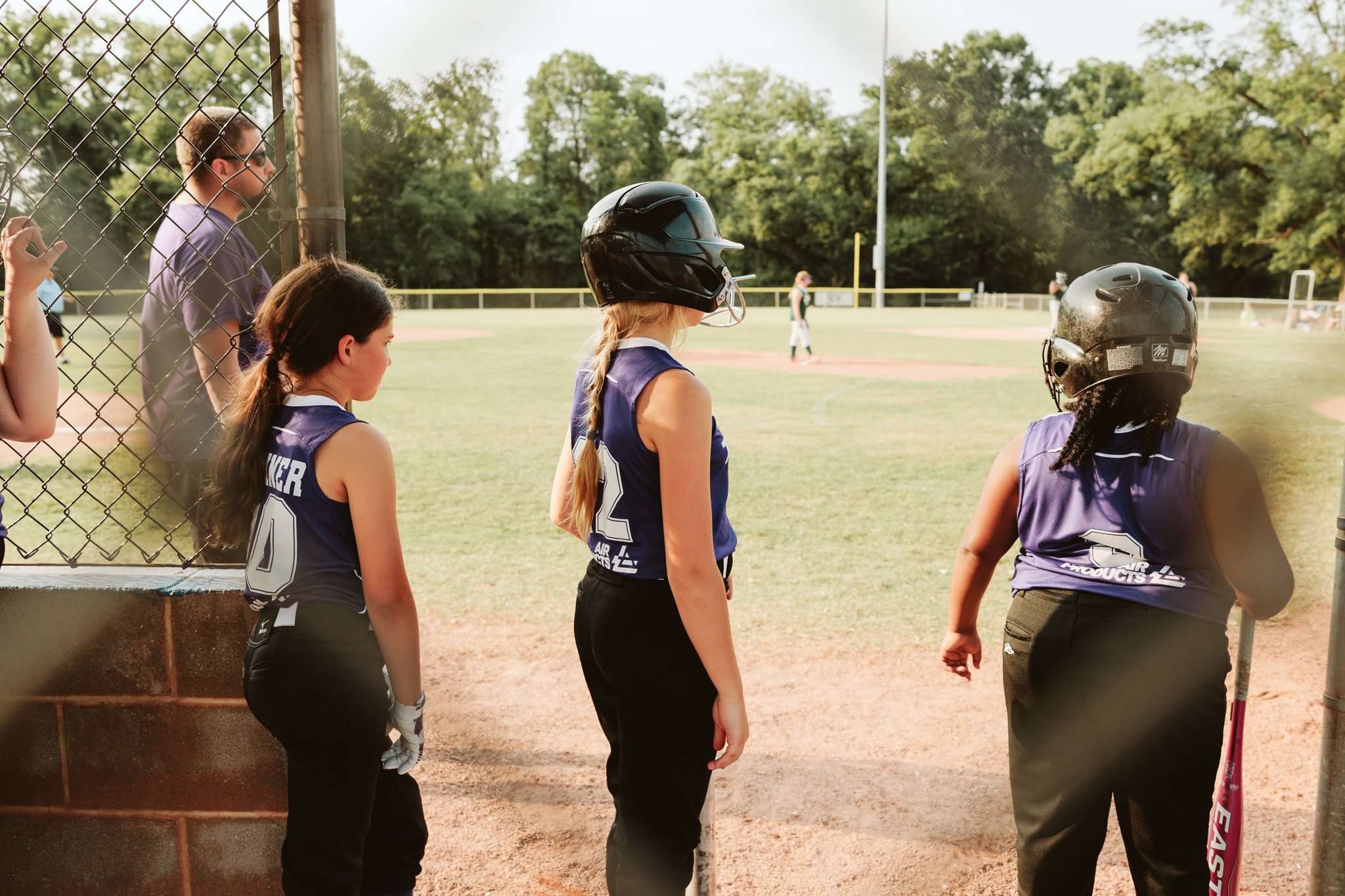 Three youth softball players wearing helmets and purple jerseys standing behind the fence watching the game at CSP.