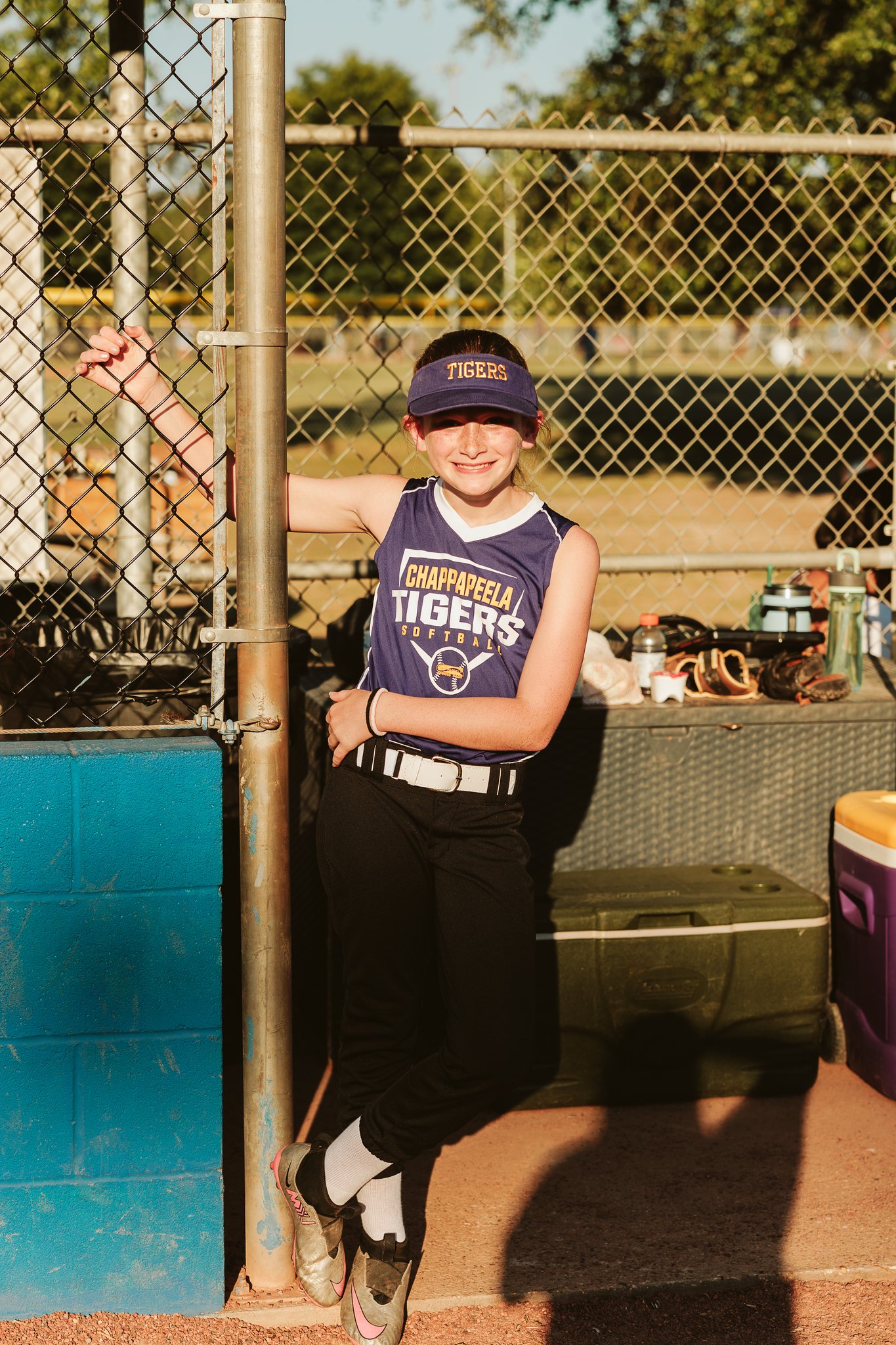 Smiling youth softball player in a purple Chappapeela Tigers jersey and visor leaning against the dugout fence at CSP.