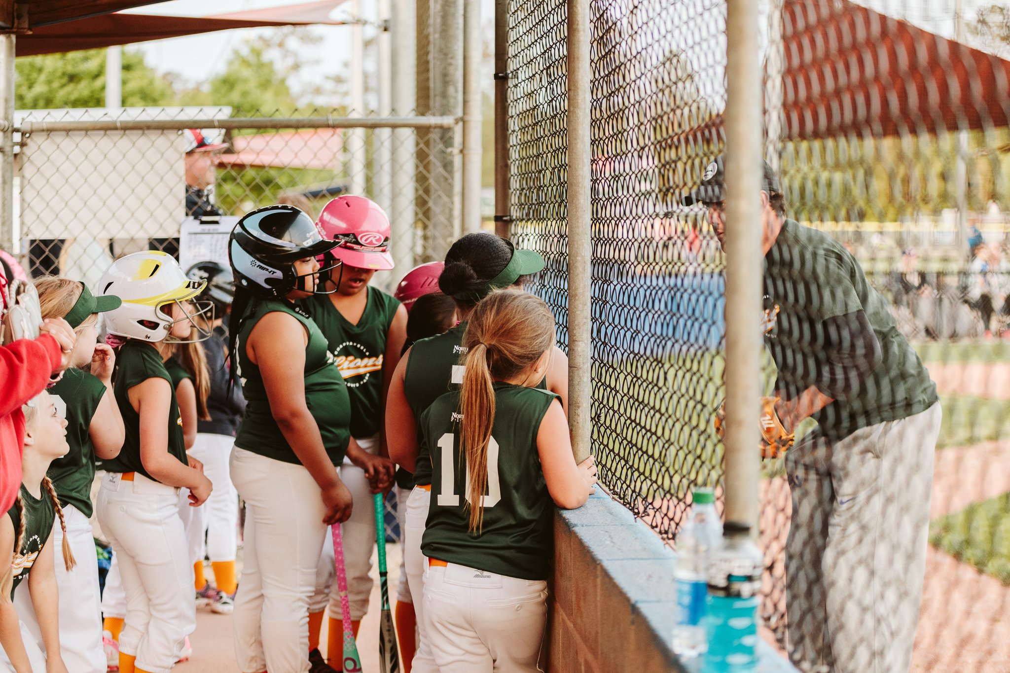 CSP softball coach giving instructions to a team of young players wearing green jerseys and helmets by the dugout fence.