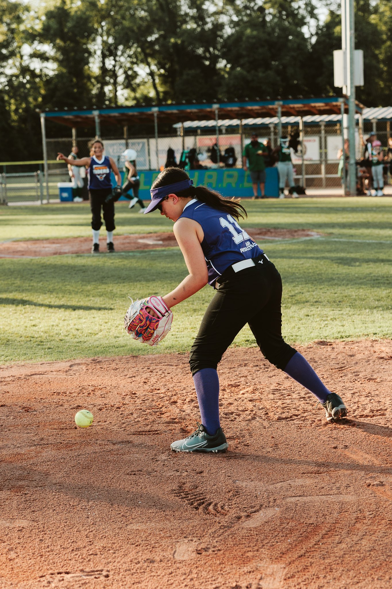 CSP youth softball player in a purple jersey bending down to field a ground ball on the dirt infield during a game.