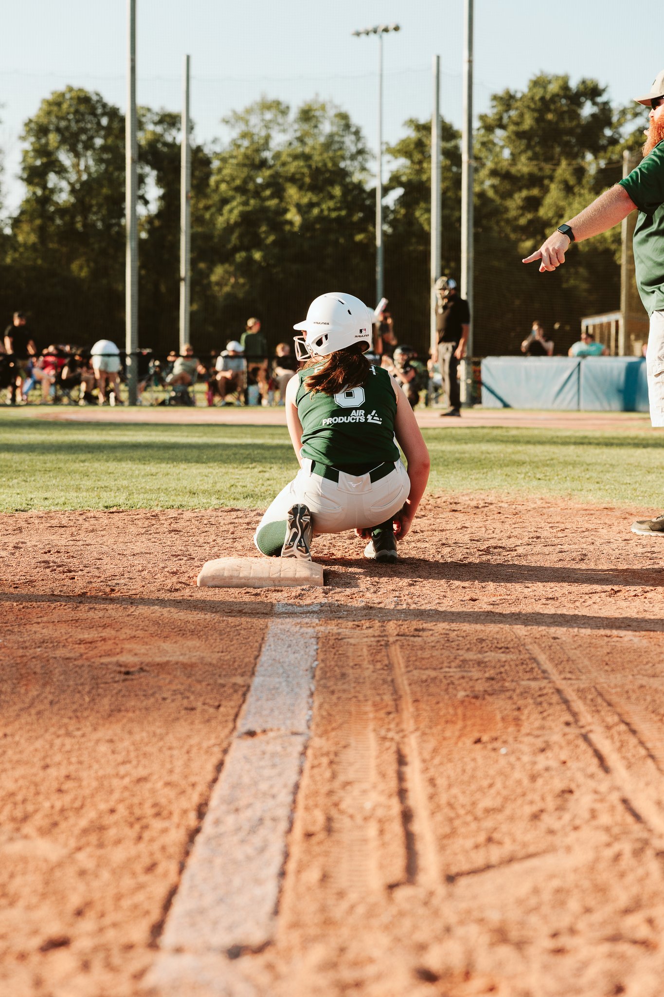 CSP youth softball player wearing a white helmet crouching on a base while a coach points towards the field during gameplay.