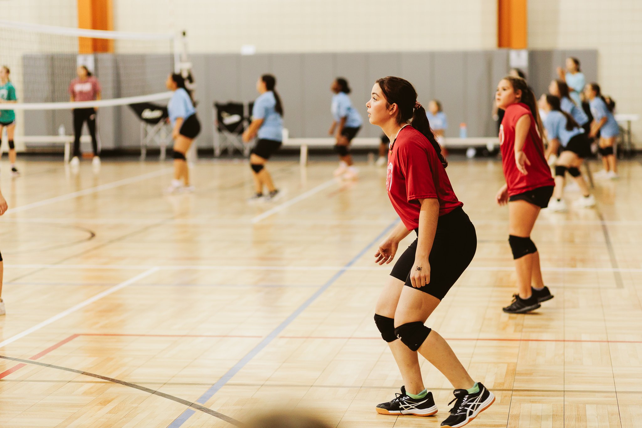 Youth volleyball player in a red jersey standing in a defensive ready position on the indoor court at Chappapeela Sports Park.