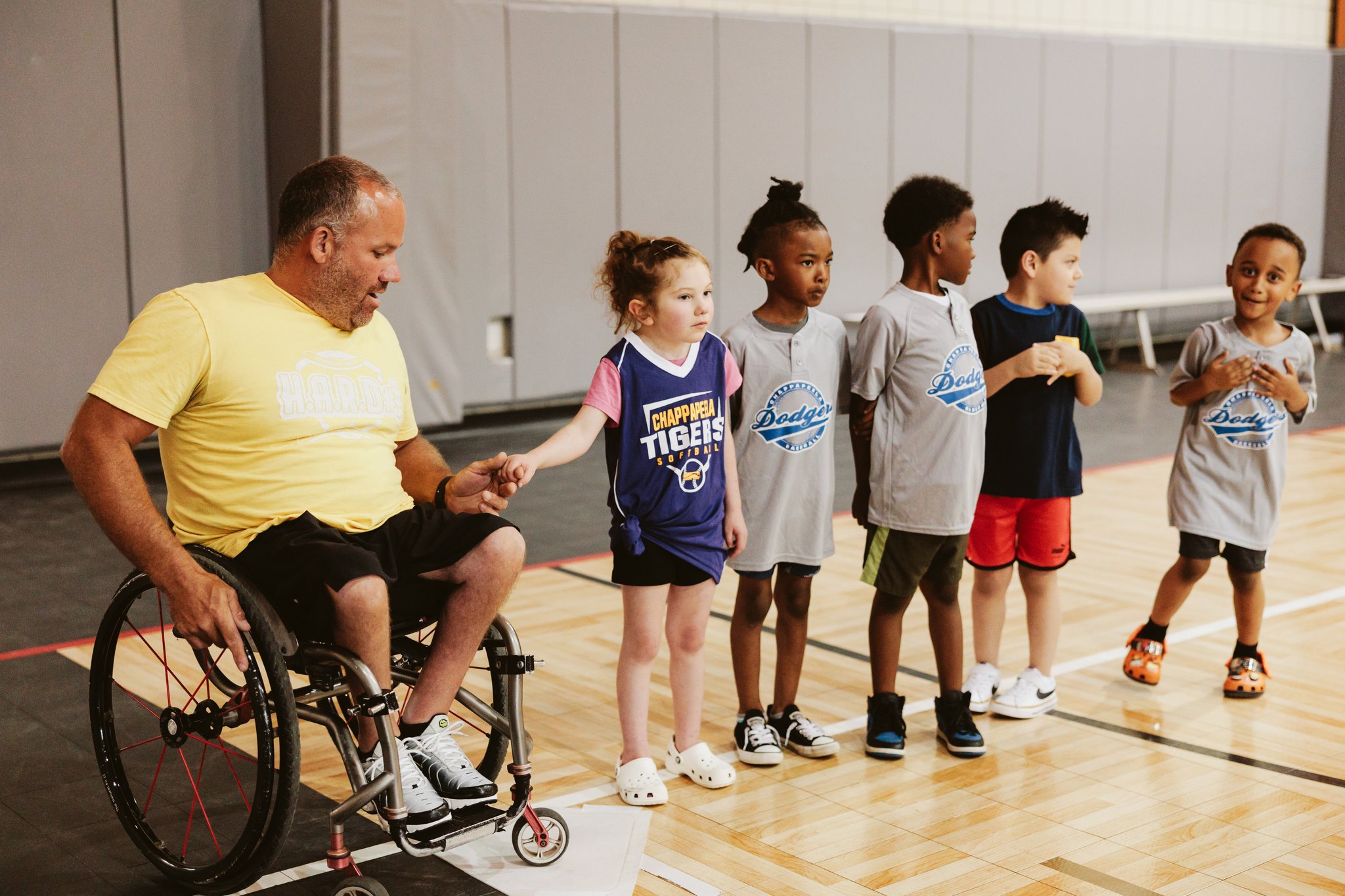 Coach in a yellow HARD #1 shirt and wheelchair fist-bumping a young participant while other children wait in line during an indoor ASPIRE adaptive sports session.