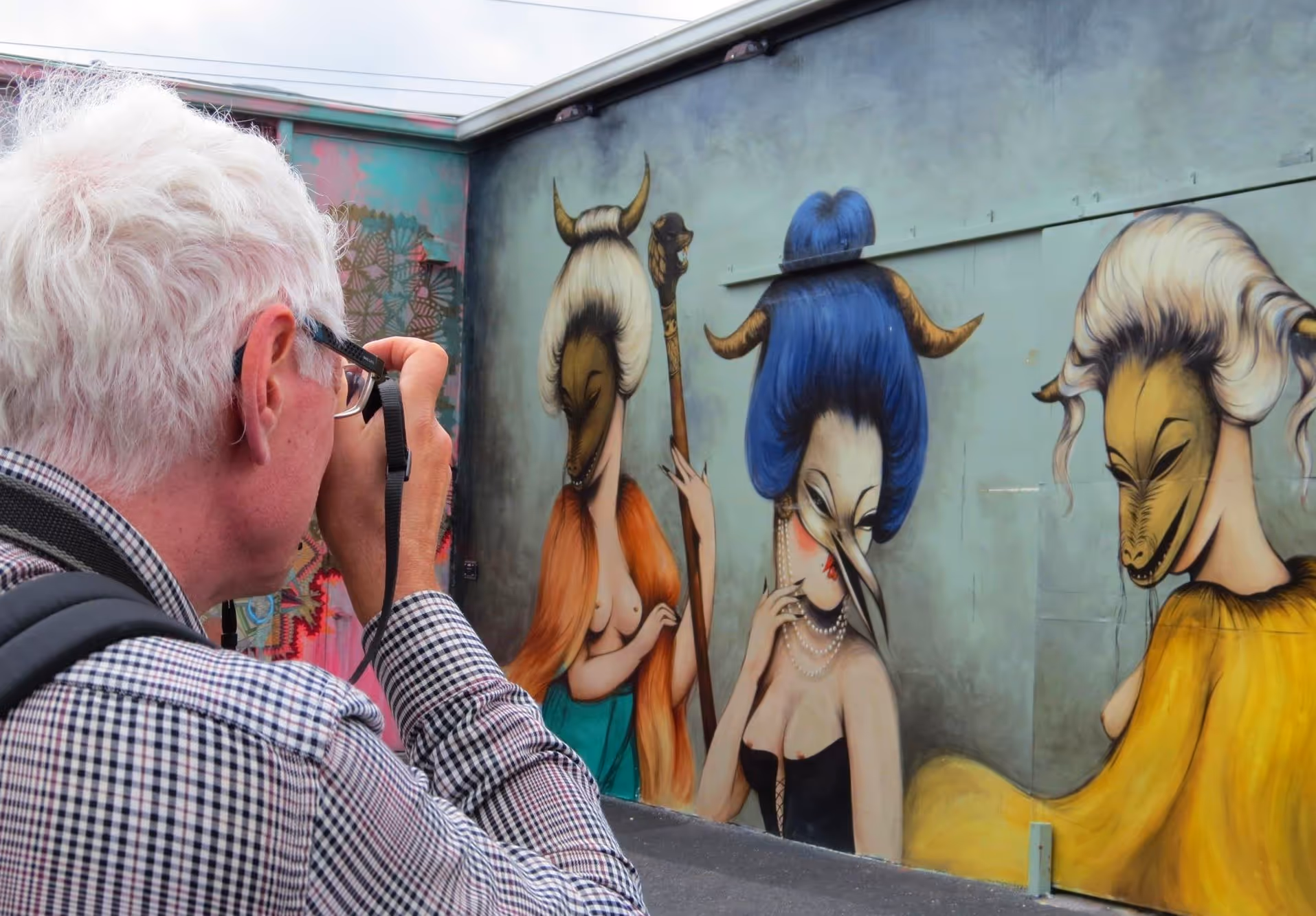 Older man taking a photo of a strange looking mural of women in animal masks