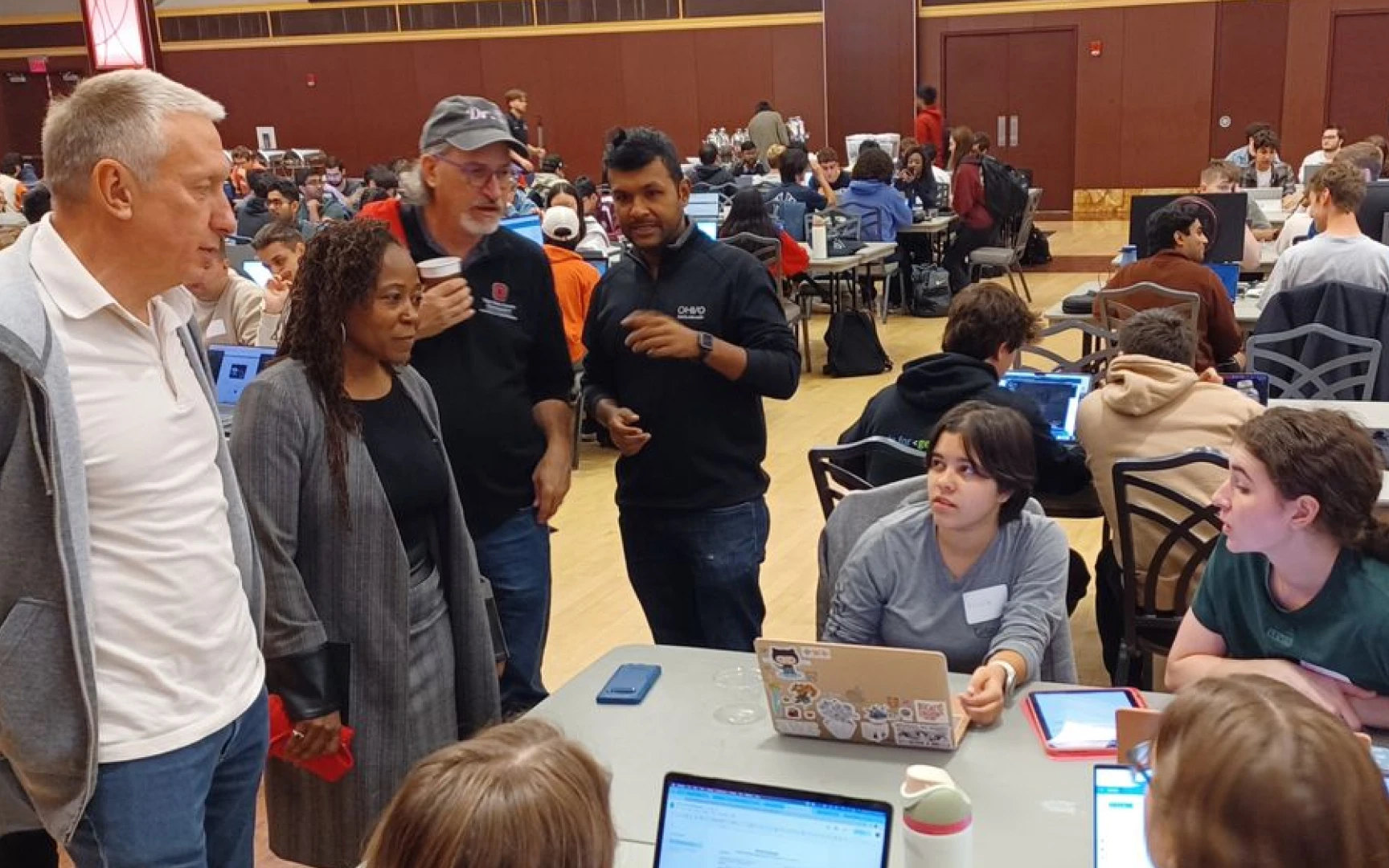 Group of adults and students engaged in conversation and working on laptops in a large, crowded classroom or workshop setting.