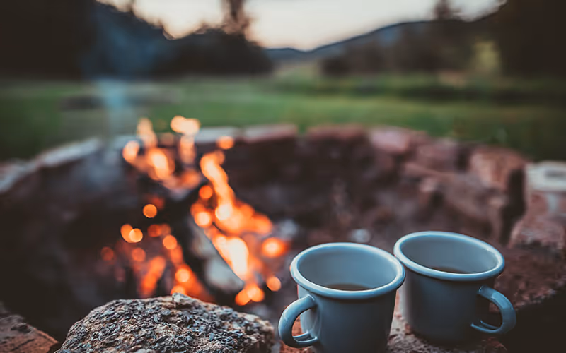 Two cups of coffee sitting on a rock in front of a fire.