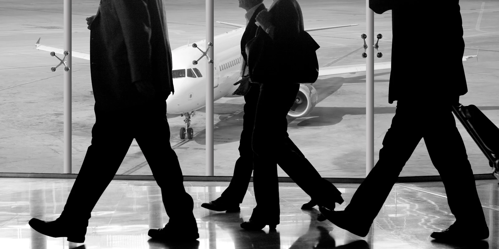 Business travellers in suits walk through an airport terminal with their luggage as a plane waits at the gate