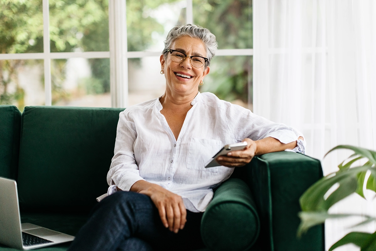 Smiling woman with short gray hair and glasses, dressed in a white shirt, sitting on a dark green couch holding a smartphone.