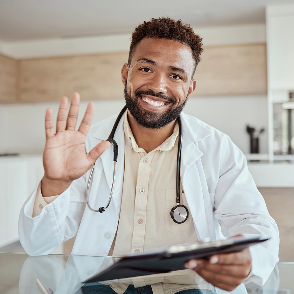 Smiling male doctor in a white coat with a stethoscope around his neck waving and holding a clipboard.