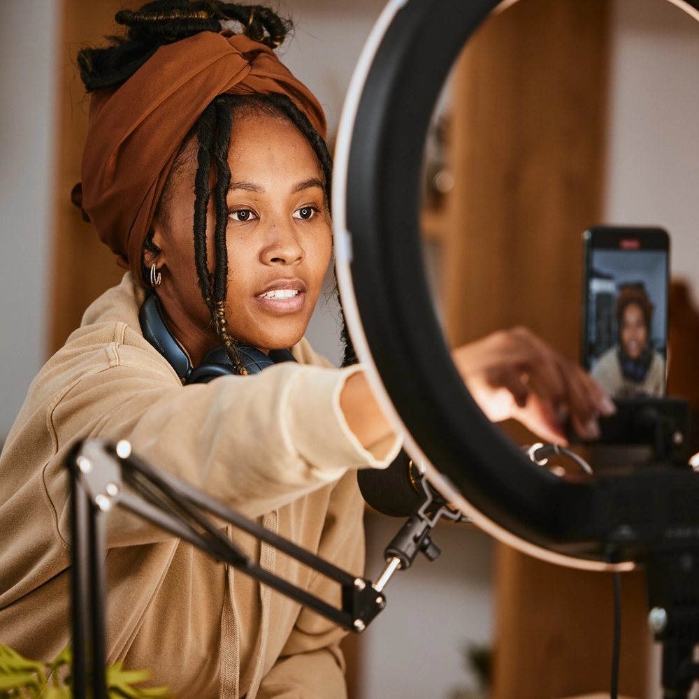 Woman with head wrap adjusting smartphone on a ring light for recording or livestreaming.