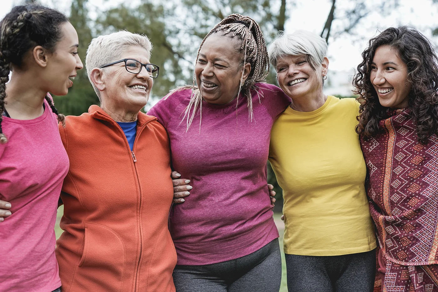 A group of diverse women standing together outdoors, laughing and linking arms while wearing colorful activewear.