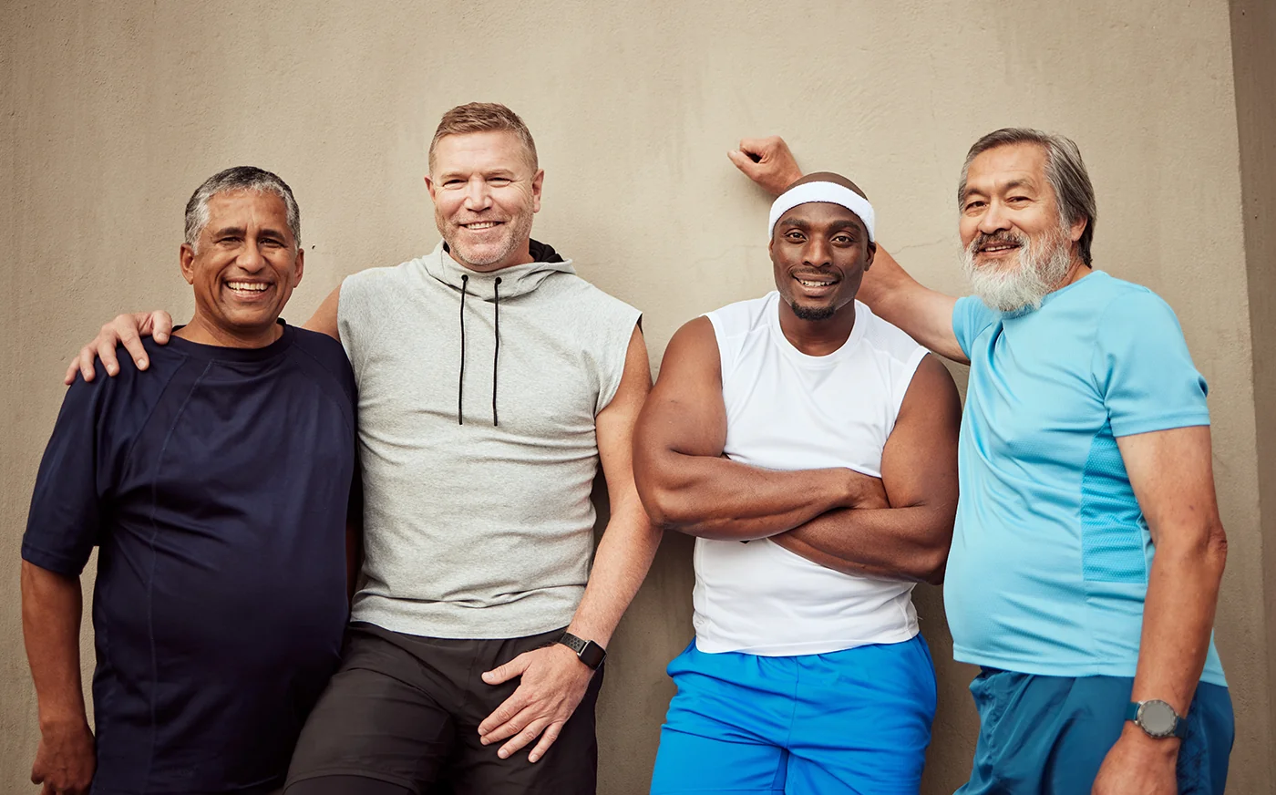 Four men of different ages and backgrounds standing against a wall, smiling after exercise and showing camaraderie.