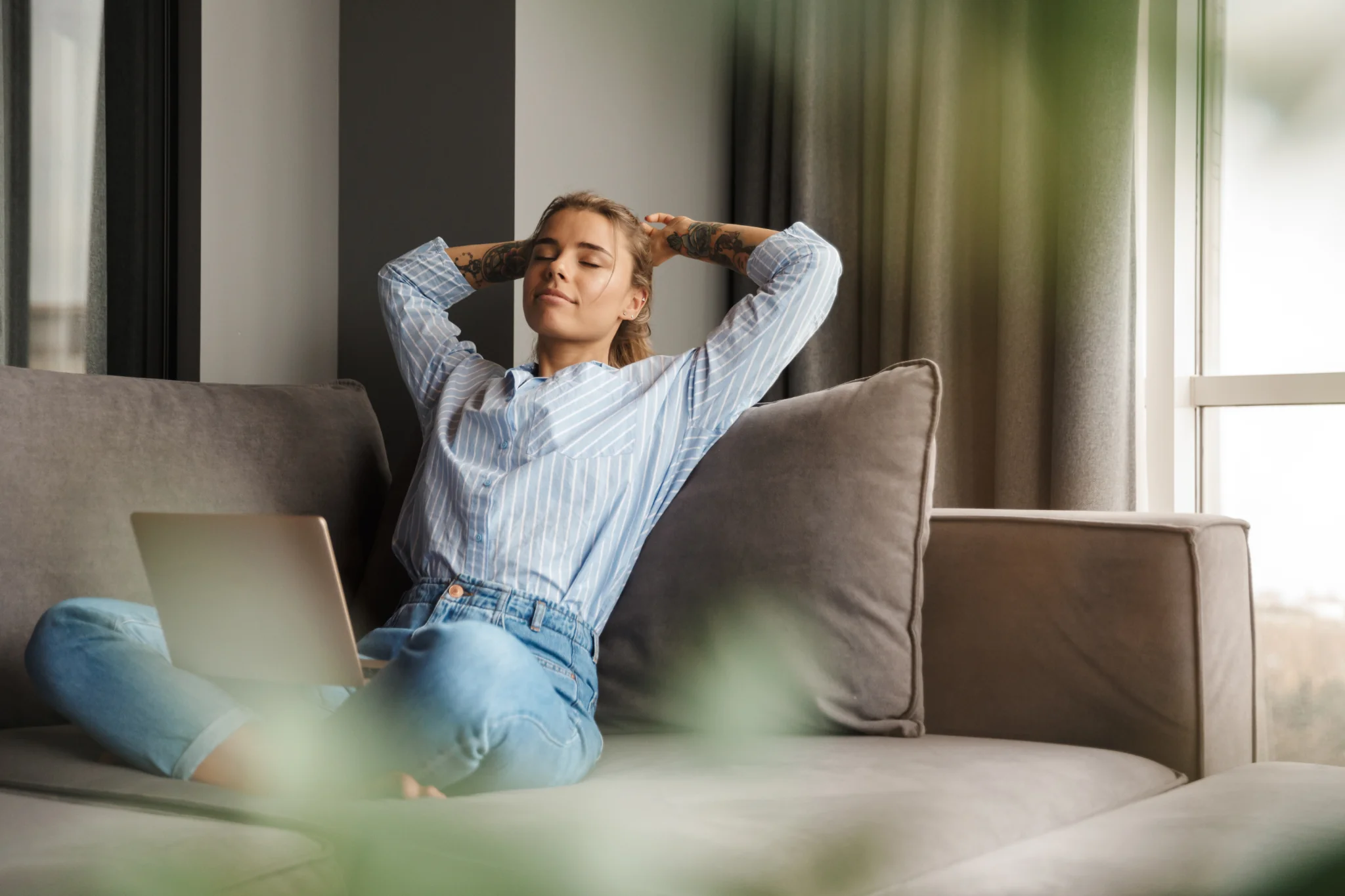 A relaxed woman sitting cross-legged on a sofa with a laptop nearby, eyes closed and arms behind her head in a calm home setting.