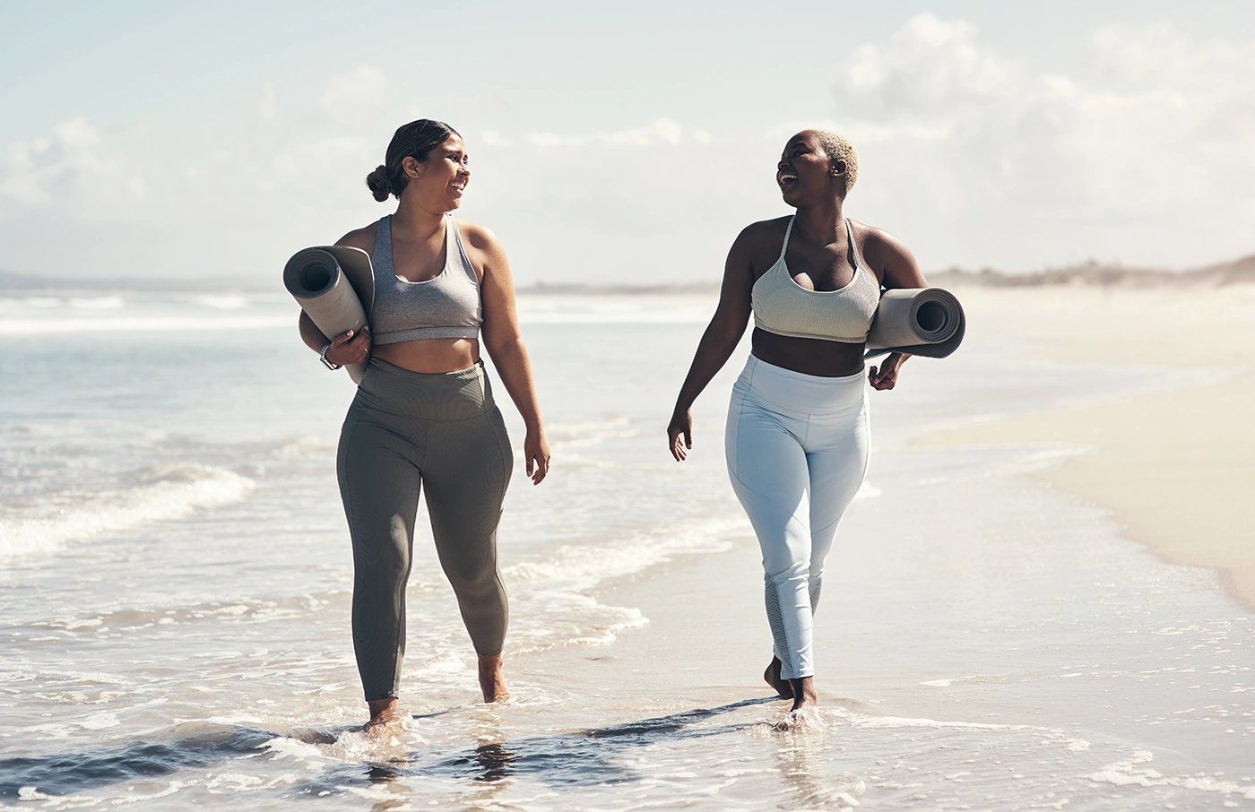 Two full-figured women walking on the beach in work out gear carrying yoga mats.