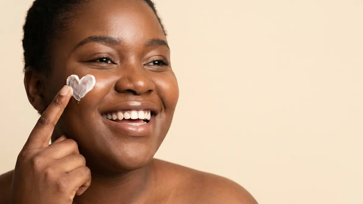 Beautiful black women draws a heart on her cheek with face cream.
