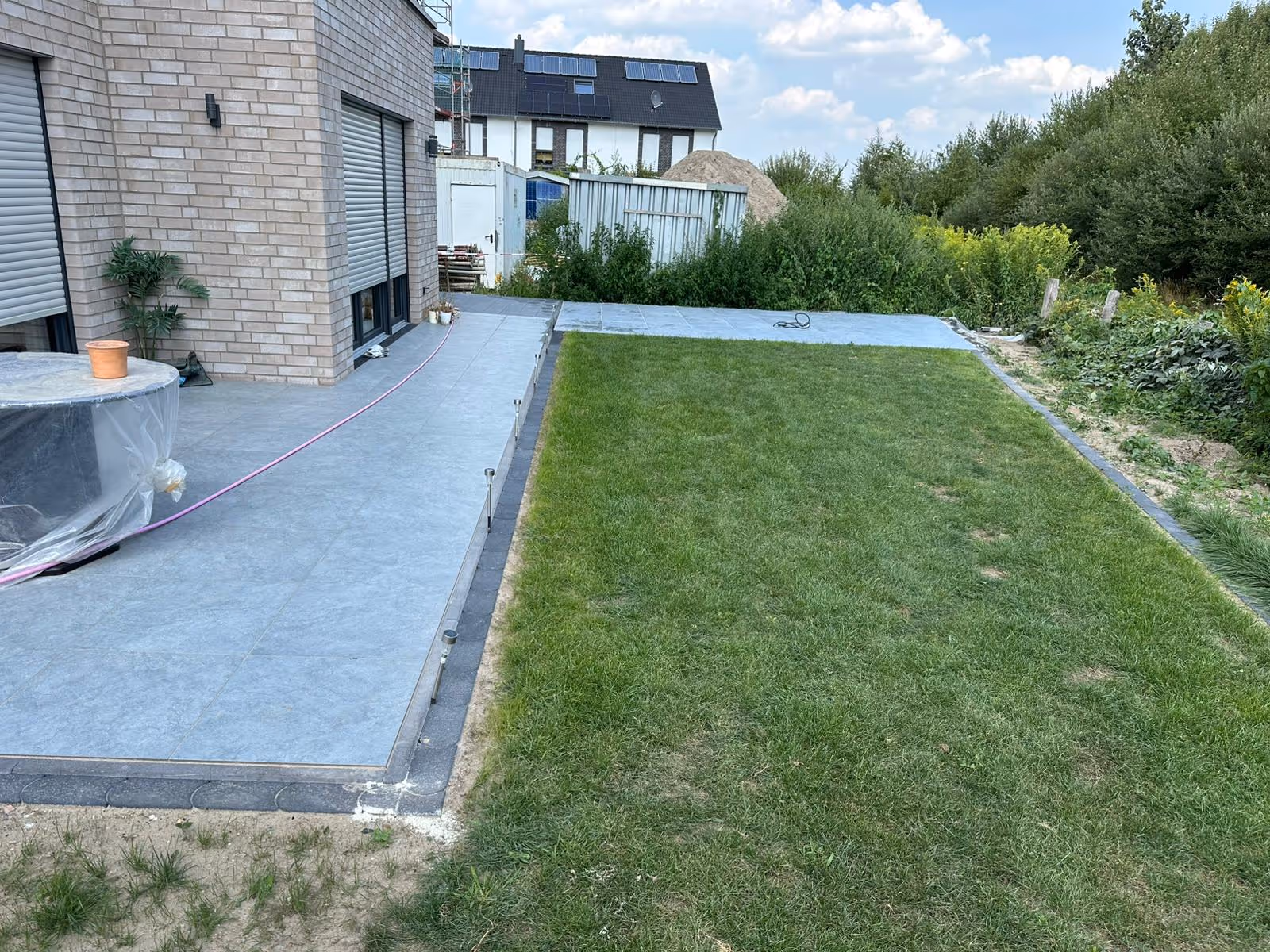 Lange, schmale Terrasse aus hellgrauen Platten mit dunkler Randeinfassung neben einer Rasenfläche - Lindemann Bau GmbH