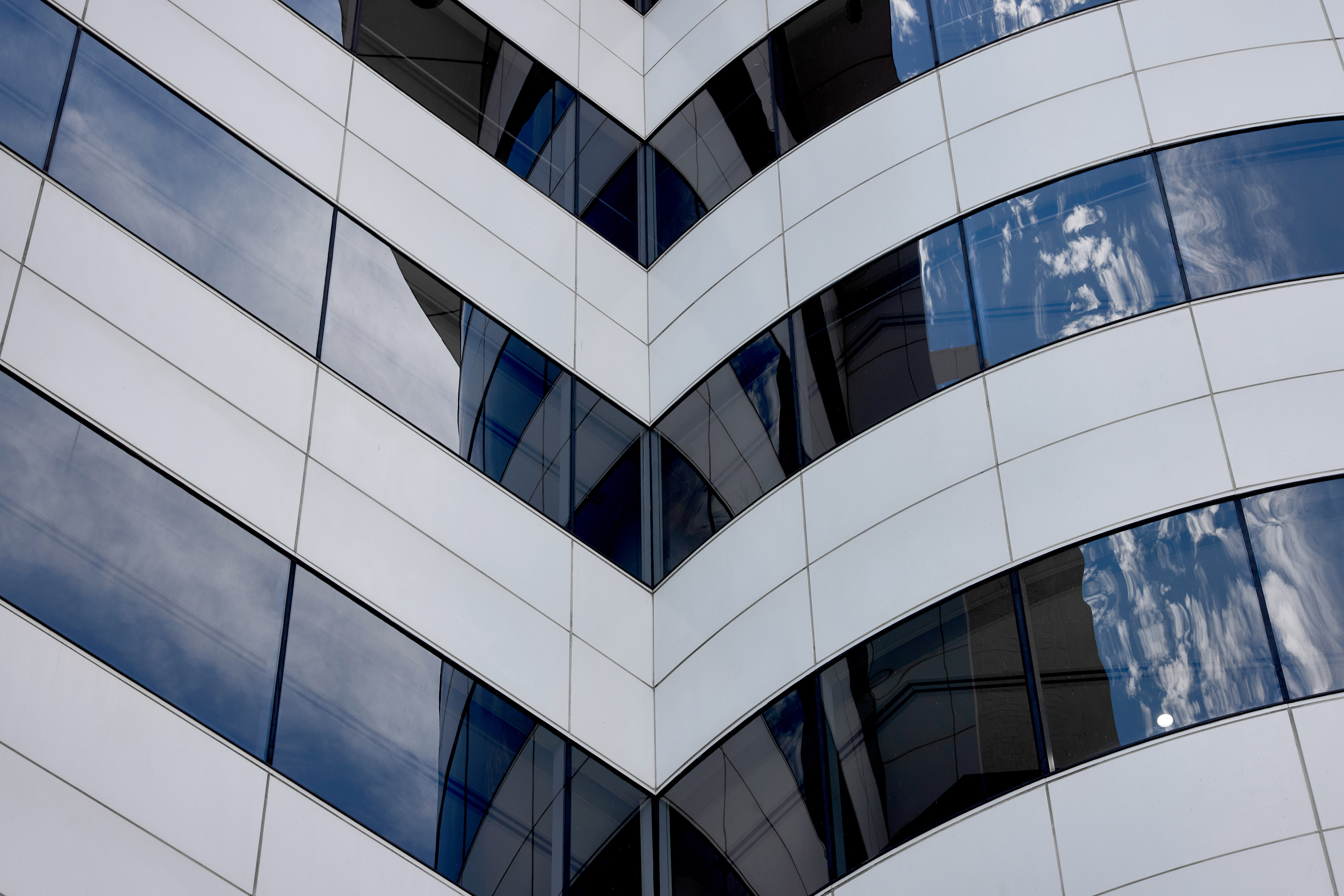 Close-up of a modern building corner with white panels and reflective blue-tinted glass windows showing sky reflections.
