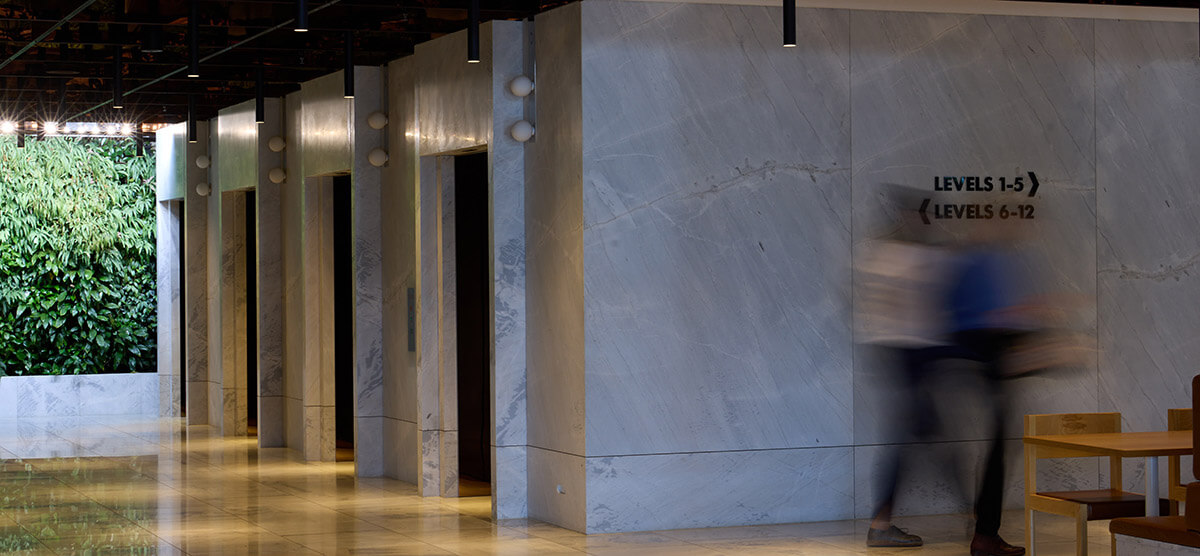 Interior hallway with marble walls, a row of elevator doors, a living green wall, and two blurred figures walking past floor level signs.