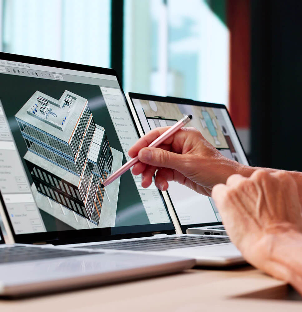 Person using a stylus to work on 3D architectural building design on a laptop screen with another laptop showing a floor plan.