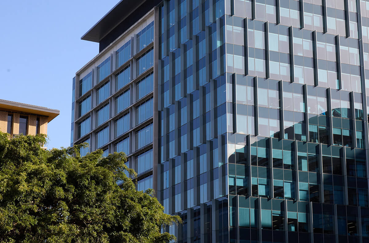 Modern glass office building with patterned facade reflecting nearby structures, next to green trees under blue sky.